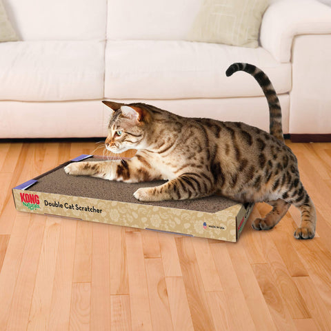 Tabby cat scratching on a KONG Naturals double cardboard scratcher on a light wood floor in front of a cream sofa.