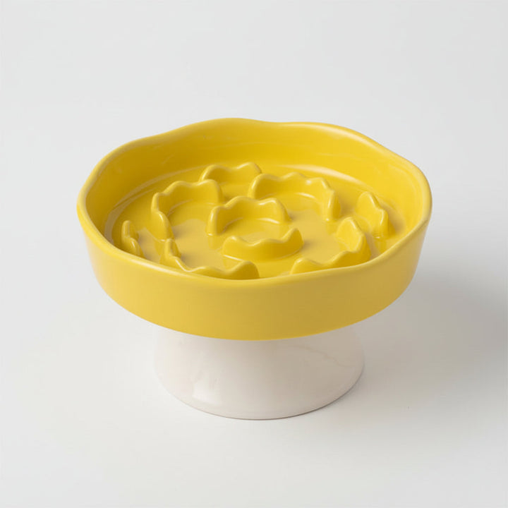 Yellow ceramic slow-feeder bowl with a raised flower-shaped center and white pedestal base, shown on a white background.