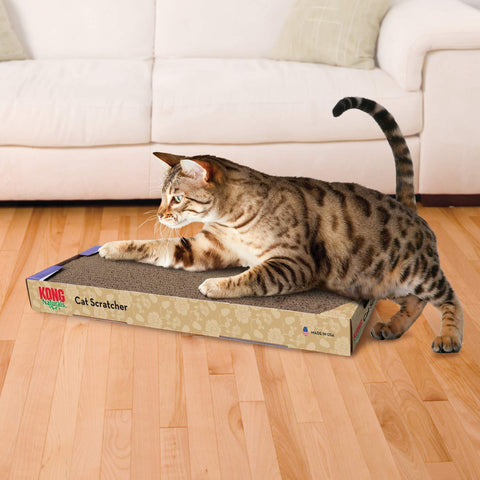 Tabby cat pawing at a KONG Naturals cardboard scratcher on a wood floor in front of a couch.
